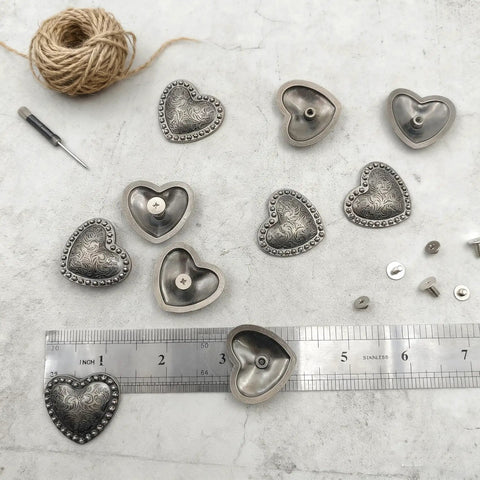 Kick Up Your Craft Game With Heart Shaped Rivets Screw Back Zinc Alloy Studs Solid Silver Metal Conchos for DIY projects arranged on a surface.