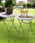 32 Inch Plastic Folding Round Bar Height Table With Gray Metal Finish For Outdoor Parties Cafes Restaurants Backyard Weddings Durable HDPE Construction 350 Lb Capacity Easy Setup White. Tables with laptop and trophies on lawn.