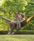 Man and child sitting in Super Comfy Double Hammock With Mosquito Net For Outdoors Portable Lightweight Nylon Parachute Hammock With 10ft Tree Straps Great For Camping Hiking Backpacking Beach And Patio Green Khaki, set up between trees.