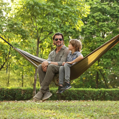 Man and child sitting in Super Comfy Double Hammock With Mosquito Net For Outdoors Portable Lightweight Nylon Parachute Hammock With 10ft Tree Straps Great For Camping Hiking Backpacking Beach And Patio Green Khaki, set up between trees.