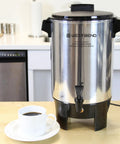Image of a 30 Cup Coffee Urn With Automatic Temperature Control And Fast Cup Per Minute Brewing Easy Cleanup In Basket Measuring Guide Ready Light Highly Polished Aluminum Silver Finish Ideal For Large Offices Events And Meetings on a kitchen counter with a white cup of coffee nearby.