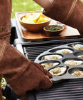 Person grilling oysters on a cast iron oyster grill pan with 12 cavities on a barbecue, showcasing nonstick surface in use.