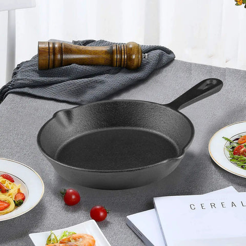 Six Inch Cast Iron Skillet With Drip Spouts For Even Heat on a table with a wooden pepper mill and plates with food