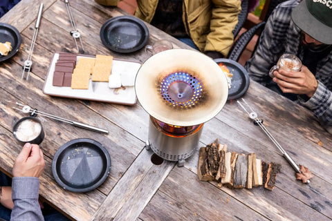 Overhead view of Mesa XL Heat Deflector with 3 detachable legs on a fire pit, surrounded by snacks and people on a wooden table.