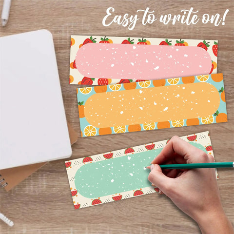 Hand writing on 30 Pcs Self Adhesive Name Stickers Set With Bright Orange Strawberry Watermelon Theme For Students Desk Tags For Classrooms Storage Box Labels Kid Friendly Classroom Decor And Supplies A03, showing various fruit-themed designs.