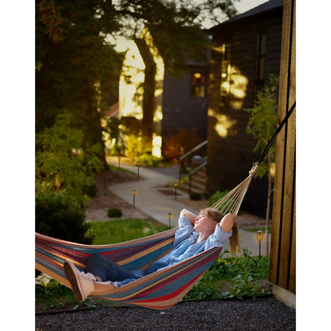 Person relaxing in hammock in garden with Outdoor Solar Flickering Torch Lights 8 Pack Waterproof Patio Tiki Lamps installed along pathway.