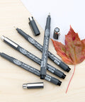 Nano Liner Drawing Pen Set Black 4 Pack Fine Point Nibs 0.1 0.2 0.3 0.5 Mm Pro Grade Ink Flow on wooden surface with leaf