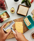 Adjustable Cheese Slicer With Four Replaceable Stainless Wires Heavy Duty Aluminum Alloy Handheld Cheese Cutter For Block And Sharp Cheeses slicing cheese on a wooden board with snacks around.