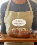 Person holding wooden board with fresh loaf of bread, wearing Sarah's Bakery apron.
