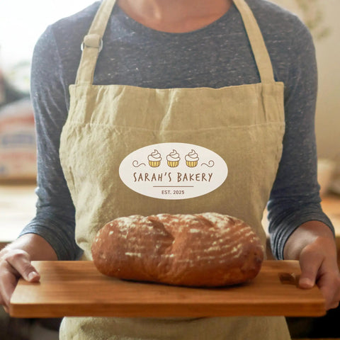 Person holding wooden board with fresh loaf of bread, wearing Sarah's Bakery apron.