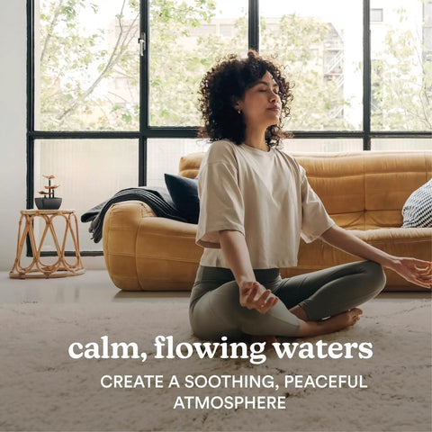 Person meditating in front of a window, seated on a carpet with a sofa and small table in a peaceful living room scene.