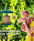 Green chew proof metal bird feeder outdoors with 3 lb seed capacity, woman holding baby next to feeding birds in garden