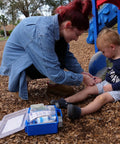 First aid kit with blue case and medical supplies opened on ground; adult bandages a child's knee at a playground.