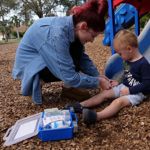 First aid kit with blue case and medical supplies opened on ground; adult bandages a child's knee at a playground.