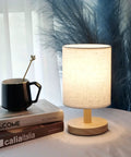 Cozy Round Table Lamp With Linen Shade, Mini Bedside Night Light on wooden base, next to a mug and books, soft glow