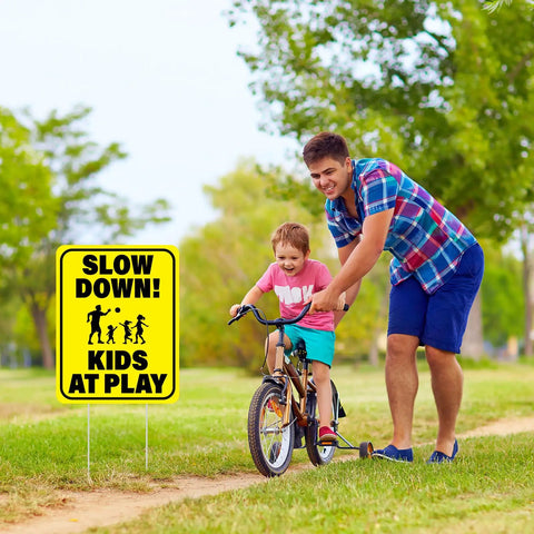 Child biking next to 3 Pack Slow Down Kids At Play Signs 14.2 By 11 Inches Double Sided Weatherproof Yard Street Safety Sign Yellow High Visibility For Neighborhoods And Outdoor Use.