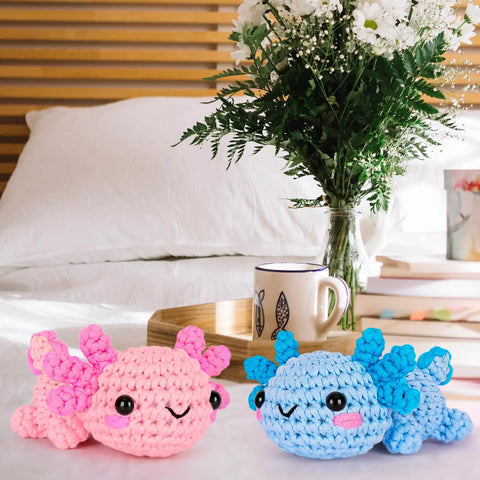 Two crocheted axolotls in pink and blue from the All In One Beginner Crochet Kit With Axolotl Pattern And Video Tutorials For Adults And Teens Starter Friendly No Fuss Do It Yourself Knitting Set With Four Easy Yarns Stuffing Hooks Safety Eyes Scissors Pattern Guide And Basic Manual on a table with flowers and a mug.