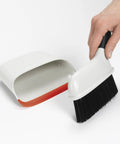 Hand using Compact Dustpan And Brush Set With Snap In Design And Bristle Cover, Soft Rubber Lip In Background