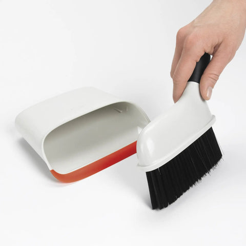 Hand using Compact Dustpan And Brush Set With Snap In Design And Bristle Cover, Soft Rubber Lip In Background