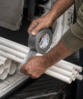 Side view of hands applying gray Duck Brand duct tape to white PVC pipes (1-pack, 60 yd).