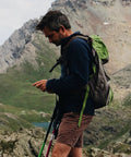 Person hiking in mountainous terrain with a green and gray backpack, using trekking poles, wearing shorts and a dark jacket.