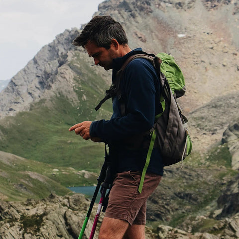 Person hiking in mountainous terrain with a green and gray backpack, using trekking poles, wearing shorts and a dark jacket.