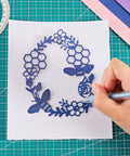 Blue laser-cut paper craft stencil featuring flowers, hexagons, and dragonflies being traced on white paper