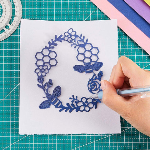 Blue laser-cut paper craft stencil featuring flowers, hexagons, and dragonflies being traced on white paper