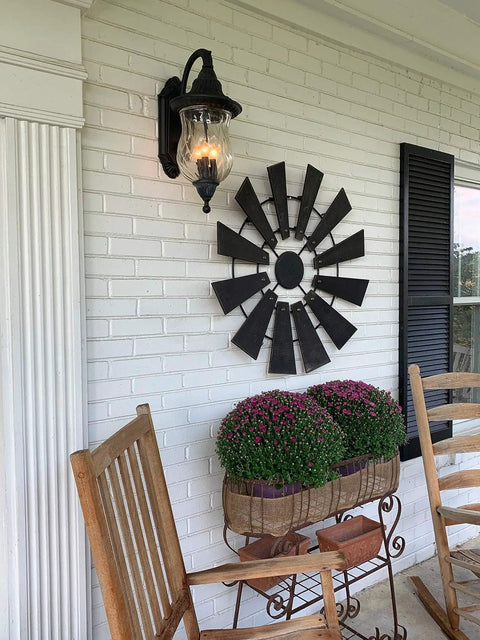Outdoor porch with rocking chair, wall-mounted black lamp, windmill decor, and flowers against a white brick wall.