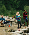 A group of friends fishing by a lakeside with camping chairs and a fire, set in a lush green forest environment.