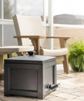 Black garden hose bin storage box outdoors, next to patio chairs, holds up to 100 ft hose, 17x17x18 in, UV fade resistant, weatherproof.
