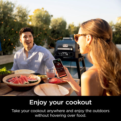Couple enjoying a meal outdoors with Woodfire Pro Connect Deluxe XL Backyard Grill and Smoker With Bluetooth App Control in background.