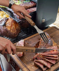 Slicing meat with a Stainless Steel BBQ Grill Tools Set With Ergonomic Wood Handles Includes Spatula Tongs Knife Fork And Carry Bag.