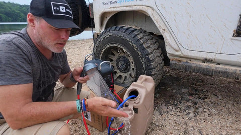 Person setting up portable propane water heater with constant temperature control for camping showers near an off-road vehicle.