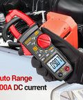 User holding All-In-One Digital Clamp Meter Multimeter For AC And DC Current And Voltage NCV Capacitance Resistance Diodes Continuity Frequency Backlit LCD 4000 Counts Handy Pro-Grade Electrician Tool For Quick Troubleshooting next to car engine