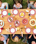 100 Count 7 Inch Red And White Checkered Paper Plates For Parties set on a picnic table with desserts and kids enjoying a gathering.