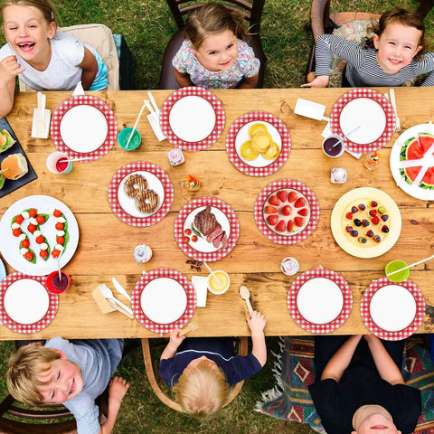 100 Count 7 Inch Red And White Checkered Paper Plates For Parties set on a picnic table with desserts and kids enjoying a gathering.