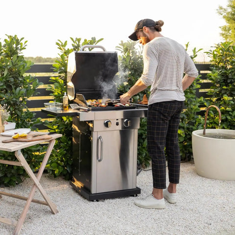 A person grilling food on a stainless steel barbecue grill in a sunny backyard area with green plants.