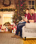 Father and son sitting by a decorated Christmas tree and fireplace with tablet, gifts and garland enhancing holiday ambiance.