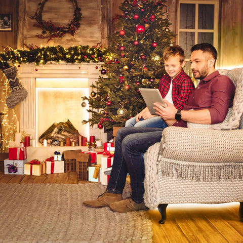 Father and son sitting by a decorated Christmas tree and fireplace with tablet, gifts and garland enhancing holiday ambiance.