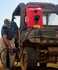 Person using Duramax Flo N Go 14 Gallon Gas Caddy On Wheels With 10 Foot Hose And Portable Fuel Tank Transfer Pump For Lawn Mowers Farm Marine And Home Use Gasoline Only Red 2024 Version in a rural setting