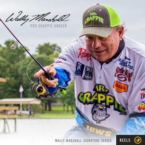 Wally Marshall holding a fishing rod, wearing a cap and branded jersey, in a lakeside setting.