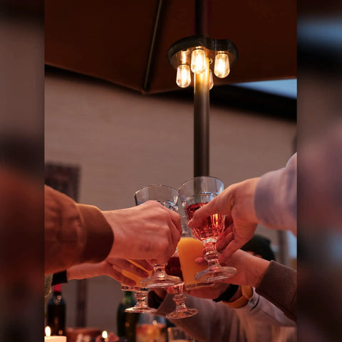 Hands toasting under an umbrella illuminated by Donut Design Patio Umbrella Light With LED Edison Bulbs Cordless Battery Powered Pole Light