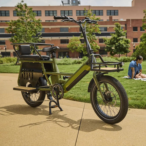 Green cargo bike with fat tires, suitable for urban commuting, parked on a walkway in a park setting with a building in the background.