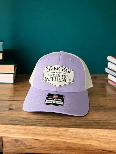 Over Par And Under The Influence Golf Trucker Hat For Women With Dimpled Ball Patch Purple Lilac Birch Color 6 Panel Structured Richardson 115 Cap Funny Golf Accessory on table.