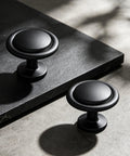 Matte Black Round Euro Style Cabinet Knobs One And One Quarter Inch 30 Pack Upgrade Your Cabinets Dresser Drawers Vanities And Coordinating With Appliances Faucets And Hardware Durable Finish And Easy Installation For Makeover Updates In Kitchen Or Bath. Two matte black round cabinet knobs on a stone surface
