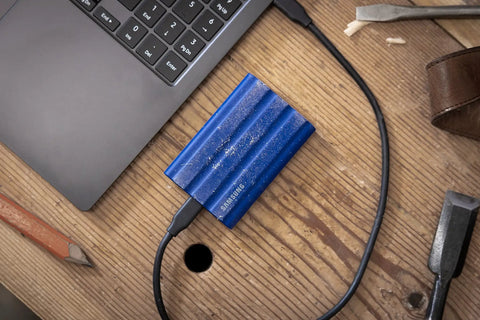 T7 Shield 1TB Portable External SSD Up To 1050 MB Per Second USB 3.2 Gen2 Rugged IP65 next to laptop on wooden desk