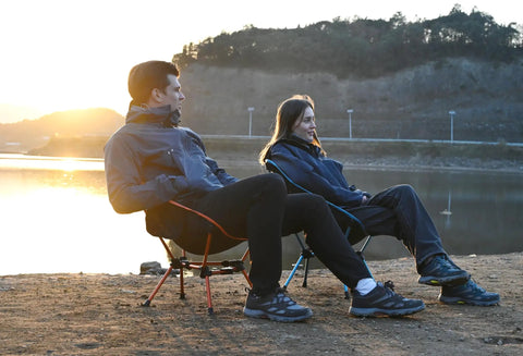 Two people sitting on Ultra Lightweight Folding Camping Chair For Outdoor Adventures And Travel, in sky blue and orange, by a lake at sunset.