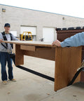 Two workers use a padded two-person Shoulder Dolly to move a long wooden desk; black straps and harnesses visible.