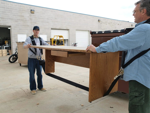 Two workers use a padded two-person Shoulder Dolly to move a long wooden desk; black straps and harnesses visible.